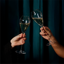 Load image into Gallery viewer, Two signature champagne flutes, filled with champagne being clinked together, in a dark & intimate setting with a blue curtain.